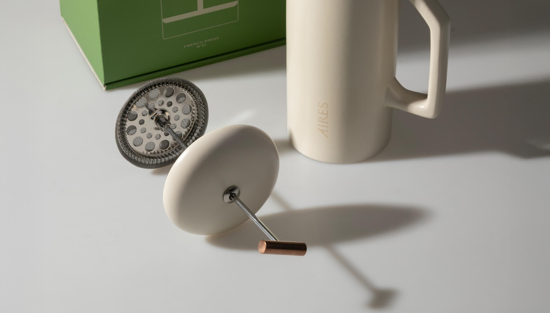 AIRES ceramic French press in tofu color with stainless steel filter placed in front of its green packaging box.